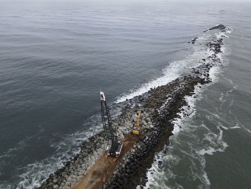 A rock structure coming out of the mouth of a river is pictured with a crane and a bulldozer on top.