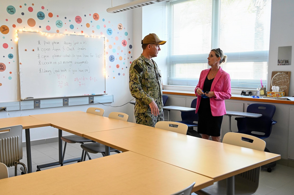 The principal of the school stands to the right explaining in a classroom to the admiral.
