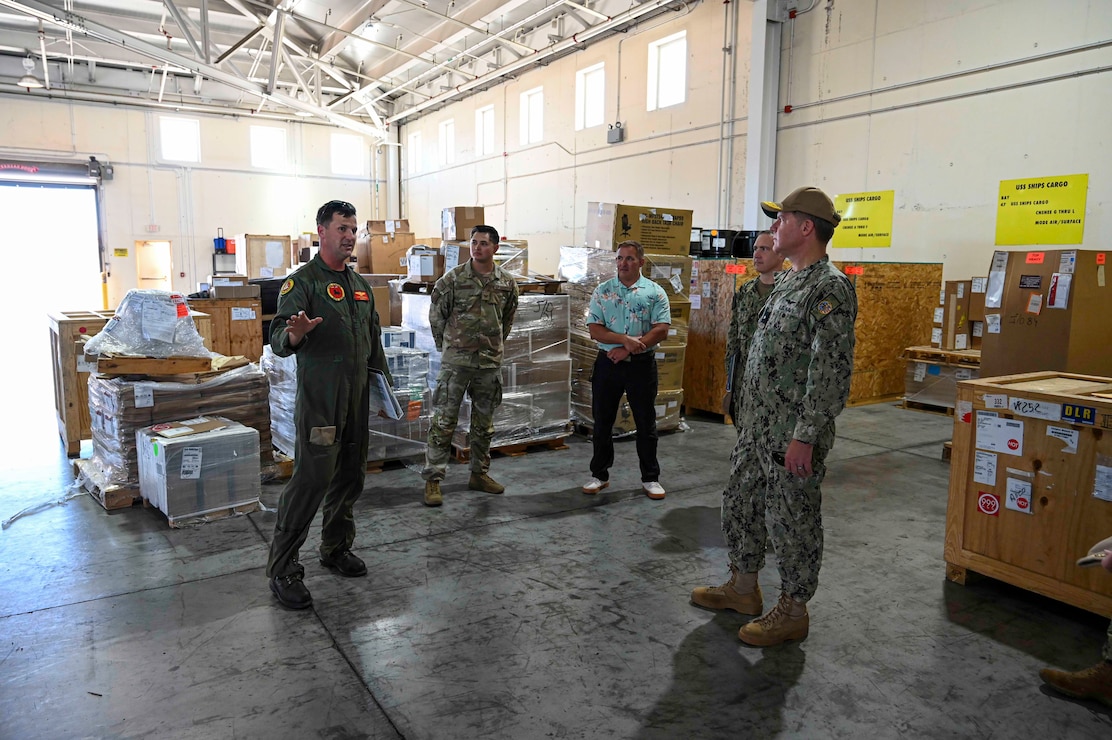 A Navy officer points at equipment in a warehouse to the Admiral.