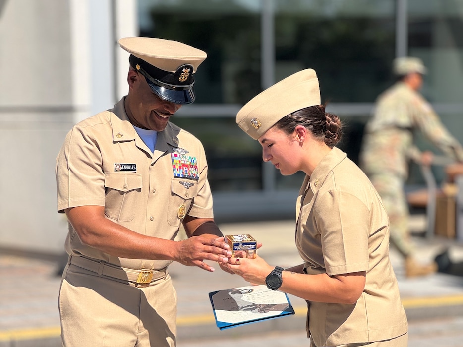 SILVER SPRING, Md. (Sept. 3, 2025) Master Chief Hospital Corpsman Phillip Jean-Gilles, command master chief (CMC) of Naval Medical Research Command (NMRC), presents a gift to Lt. Sara Tuck following a reenlistment ceremony in which Tuck administered the oath of enlistment, a career first, to Jean-Gilles. Immediately following his reenlistment, Jean-Gilles relinquished his position as CMC to Master Chief Hospital Corpsman Christopher Cariss during a change of office ceremony. NMRC, headquarters of Navy Medicine Research & Development, is engaged in a broad spectrum of activity from basic science in the laboratory to field studies in austere and remote areas of the world to investigations in operational environments. In support of Navy, Marine Corps and joint U.S. warfighter health, readiness and lethality, researchers study infectious diseases, biological warfare detection and defense, combat casualty care, environmental health concerns, aerospace and undersea medicine, operational mission support and epidemiology. (U.S. Navy photo by Tommy Lamkin/Released)