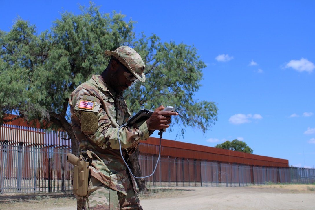 A guardsmen stands outside near a fence and a large tree and operates drone equipment under blue skies.