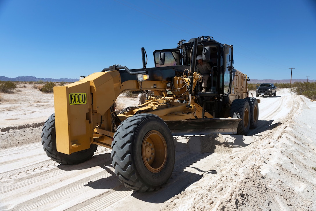 A Marine drives a large yellow construction vehicle over a sandy road under blue skies.