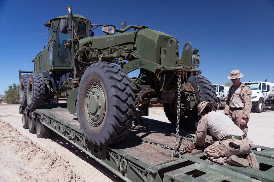 A Marine crouches in front of a large tractor parked on the bed of a truck while another Marine stands to their right and watches under blue skies.