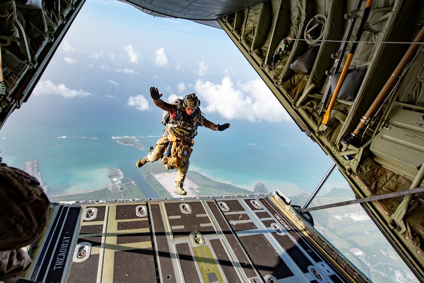 A man wearing a camouflage military uniform and parachute equipment jumps from a military aircraft. The rear of the aircraft is in front of him, and behind him is a blue sky with clouds.