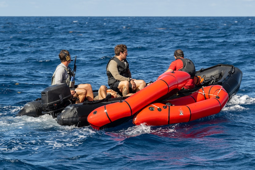 Three people sit in an orange and black life raft in the ocean.