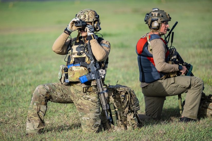 Two people wearing camouflage military uniforms kneel in a grassy field facing opposite directions. Both have weapons, and one is holding binoculars.