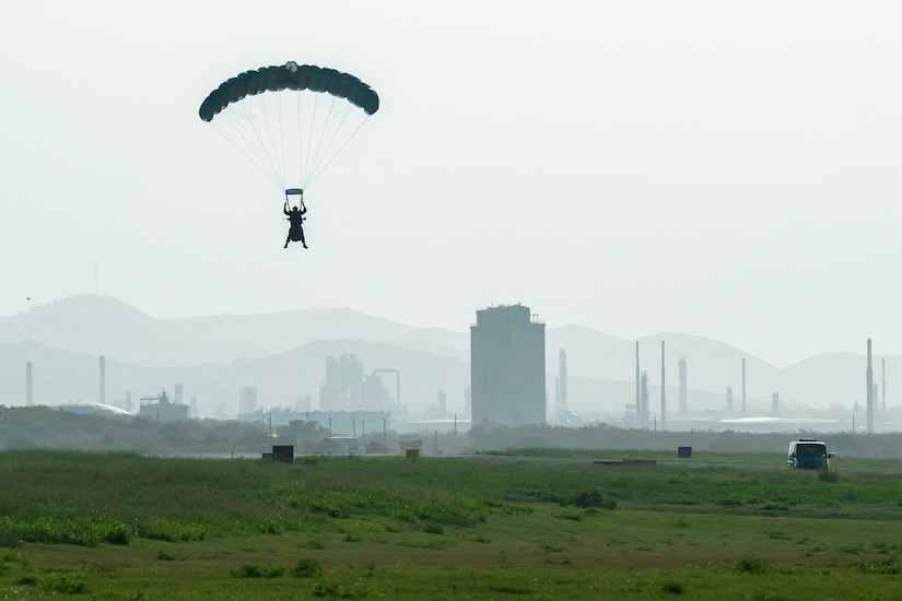 The silhouette of a person hanging from a parachute and landing in a green grassy field with buildings and mountains in the background.