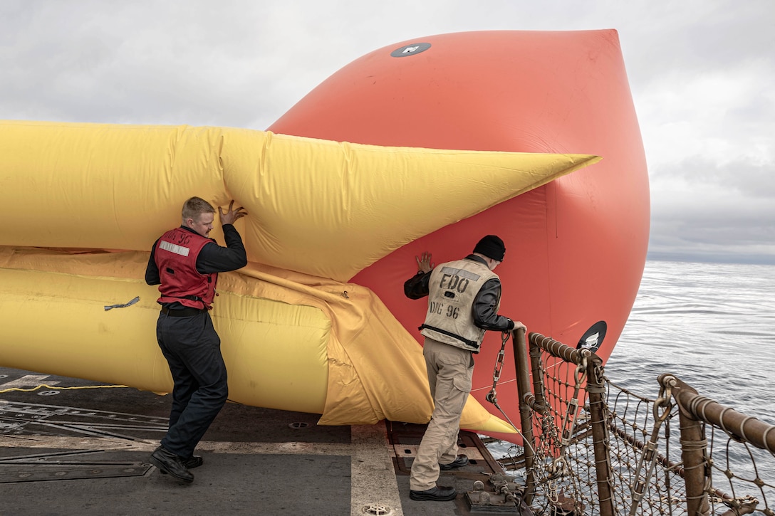 Two sailors move a large orange and yellow inflatable target from a ship into a body of water on a gloomy day.