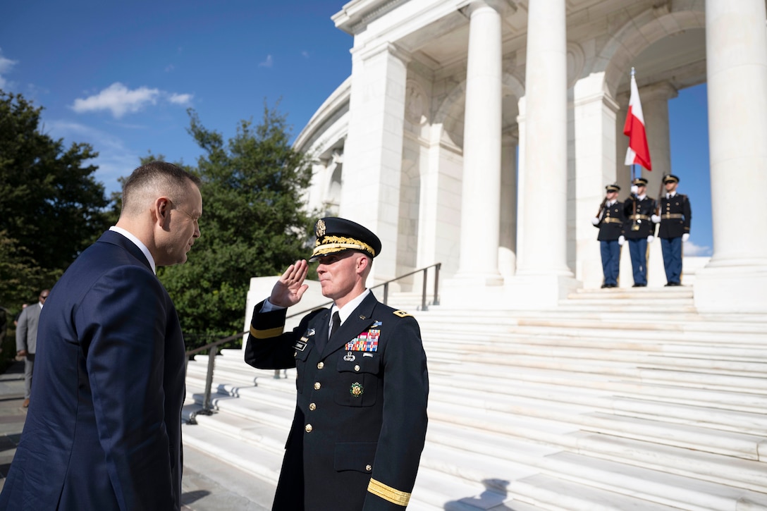 A U.S. soldier salutes the Polish president in front of a monument as three fellow soldiers stand in formation carrying rifles and a flag.