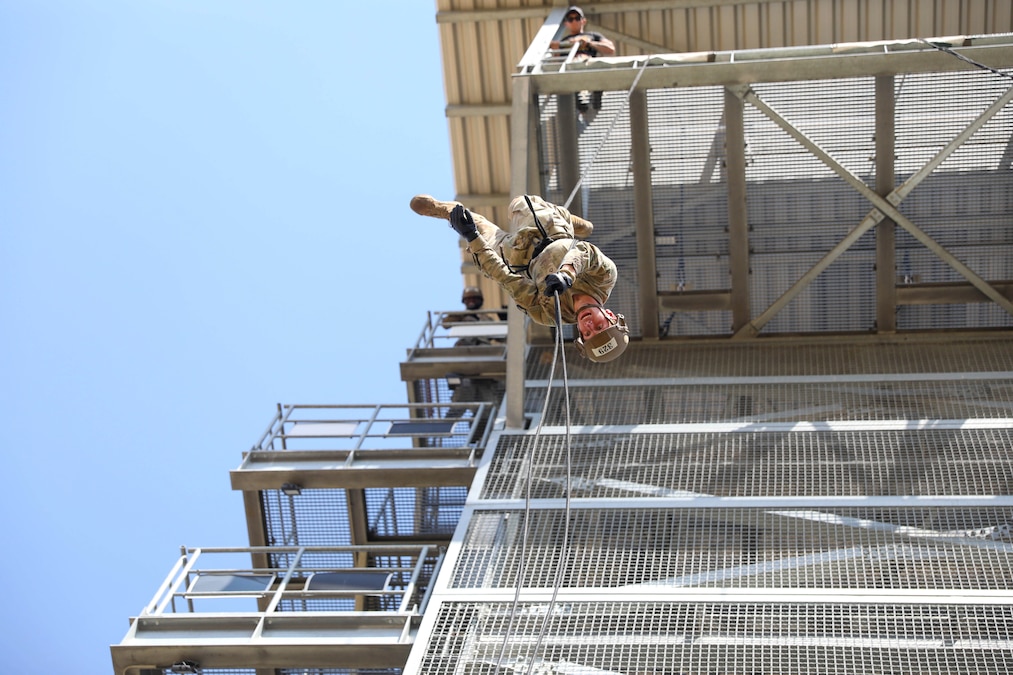 A soldier in protective gear rappels upside down from a tower as a fellow soldier watches from above.