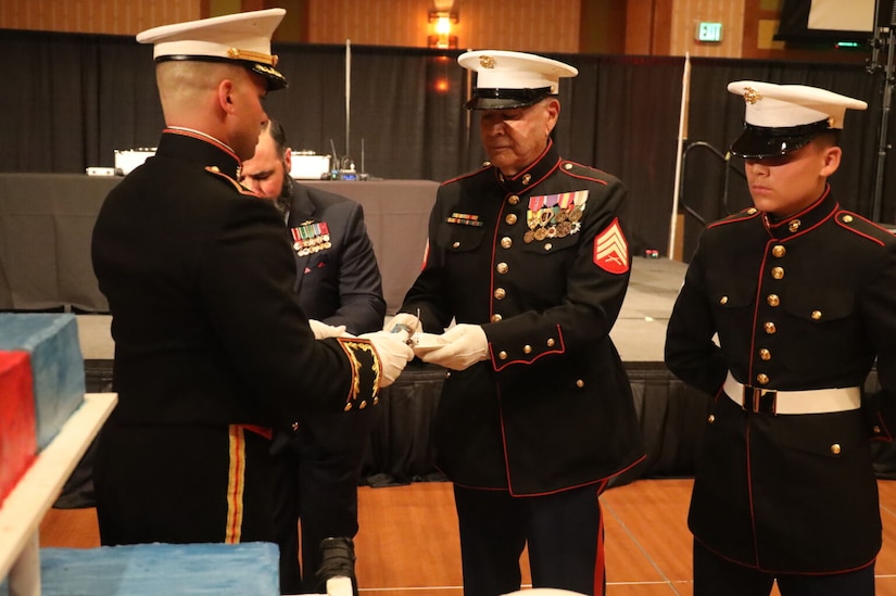 A Marine in a military dress uniform hands a slice of cake to another Marine in similar attire. There is another Marine in similar attire standing to the right.