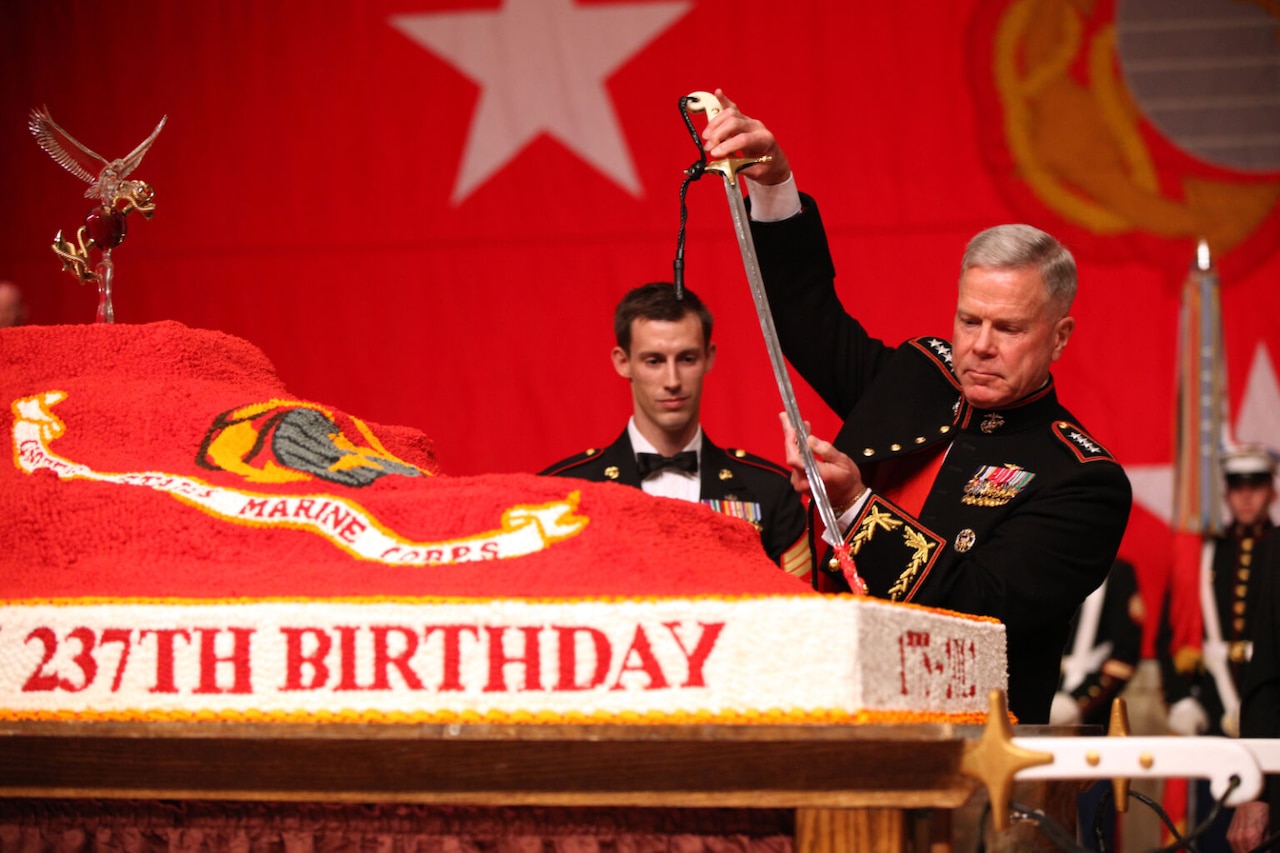A Marine in dress military attire slices into a large cake with a sword.