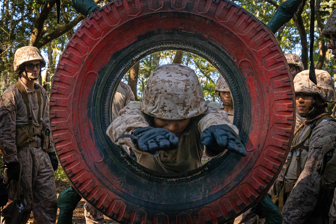 A Marine recruit maneuvers, hands first, through a red tire while other recruits stand behind and watch with a forest behind.
