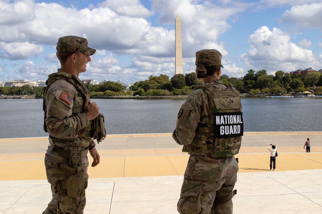 Two guardsmen in camouflage look to the right with a body of water and the Washington monument in the background, as seen from behind.