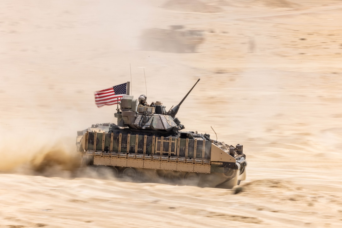 Soldiers sit on top of a moving tank with an American flag as it drives and kicks up sand during the day.