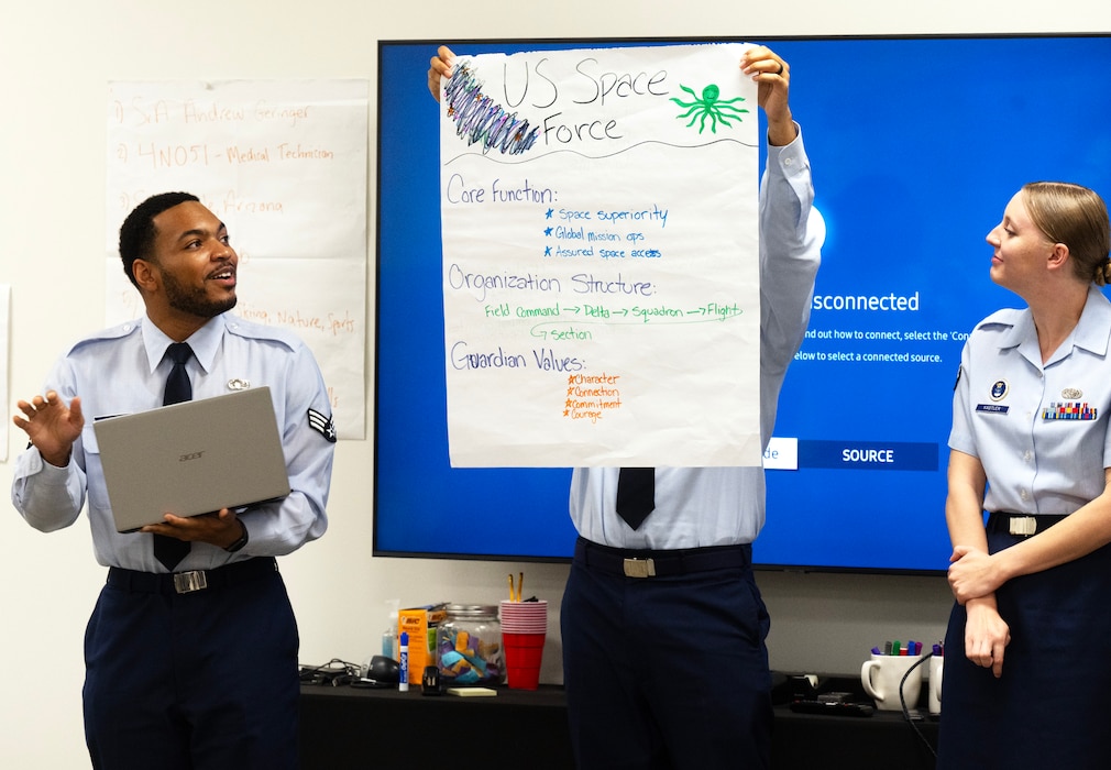 A man standing on the left holds a laptop computer in one and and gestures with his other hand as he speaks. On the right is a woman in the blue Air Force uniform. They are both looking at a large piece of white paper with information about the U.S. Space Force on it that is being held up by a third person who is hidden by the paper.