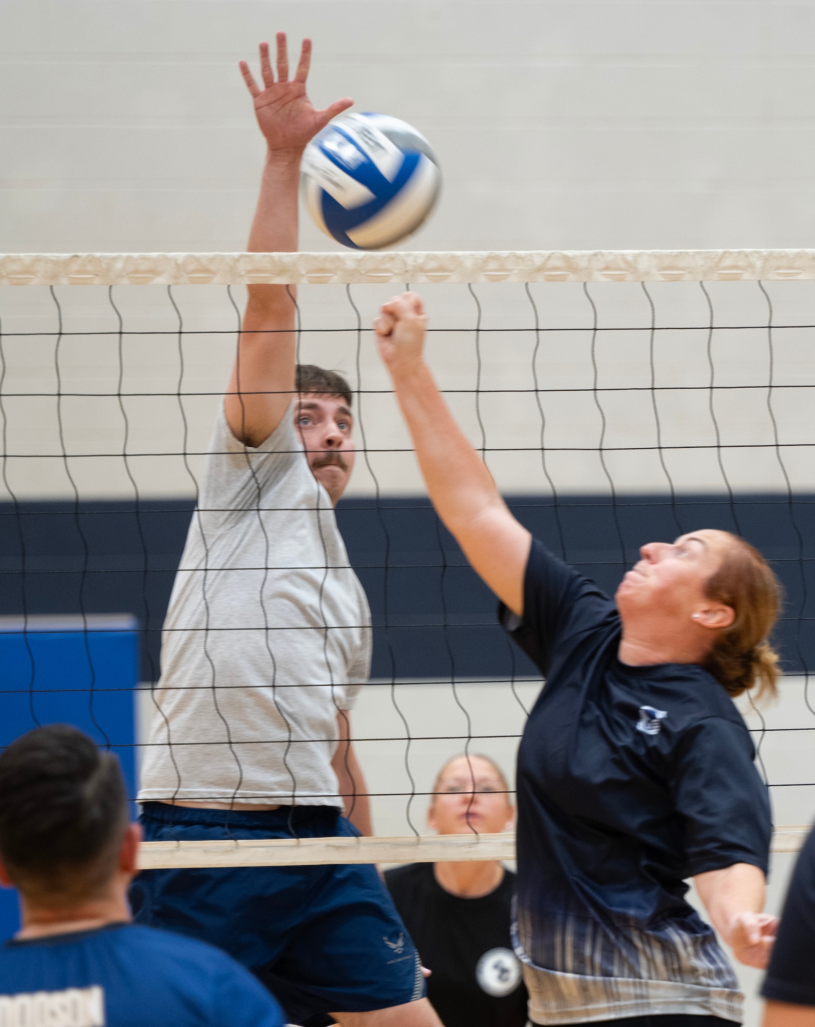 A man on the far side of a volleyball net jumps with his left hand extended as a woman on the near side of the net tries to hit the ball over.
