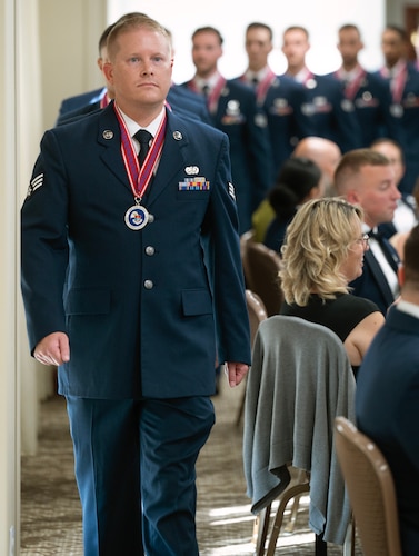 A man in a dress uniform, wearing a medallion, marches into a dining room in the front of a line of Airmen.