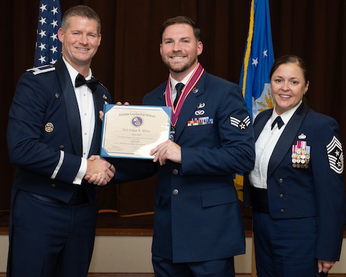 A man in formal, dress uniform stands on the left. in the center is a younger man in less formal uniform, they hold a diploma and shake hands. A woman in formal, dress uniform stands on the right.