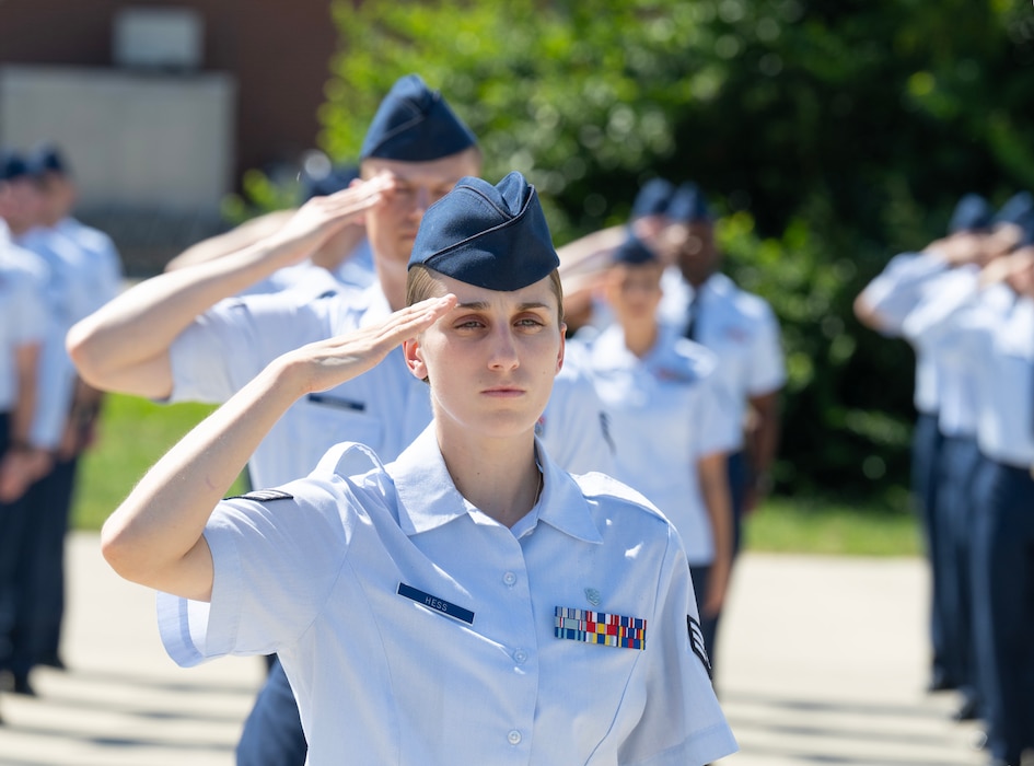 A woman in an Air Force uniform stands at attention and salutes. A formation of Airmen saluting is behind her.