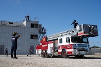 firefighter looks at the firetruck and adjusts his helmet as another firefighter exits the ladder on the firetruck.