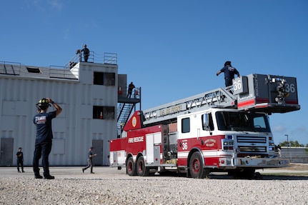 firefighter looks at the firetruck and adjusts his helmet as another firefighter exits the ladder on the firetruck.