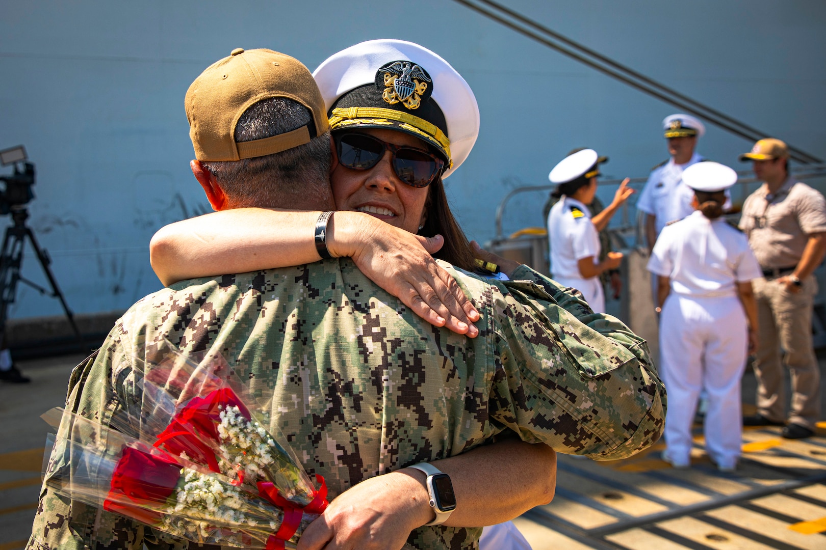 Uniformed service members hug and hold flowers standing in front of ship.