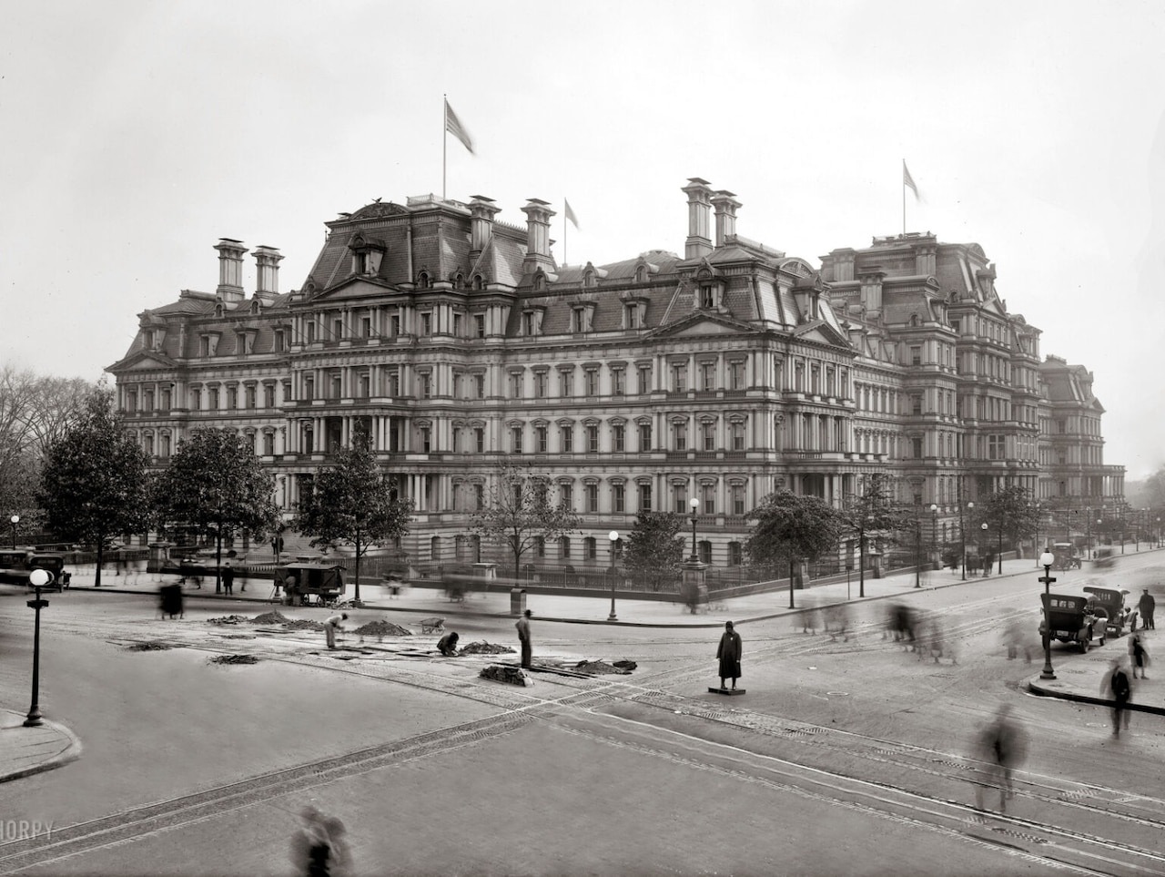 A black and white photograph of a gothic-style, five-story building with antique cars parked nearby.