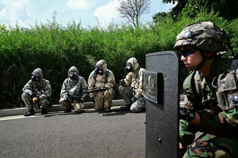 A soldier wearing a camouflage military uniform holds a protective shield in front of their body while crouching on the ground. There are four soldiers in military camouflage uniforms and gas masks crouched un the background.