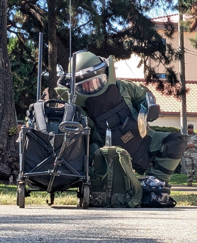 A soldier in a protective bomb suit crouches on the ground next to a backpack and roller cart. There are trees and a building in the background.