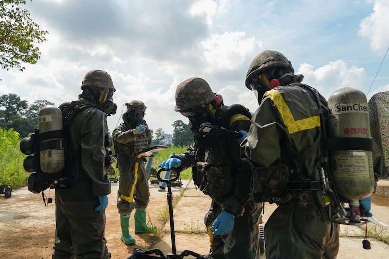 Three soldiers wearing camouflage military uniforms and personal protective equipment stand together as another soldier in similar attire in the background motions to them.