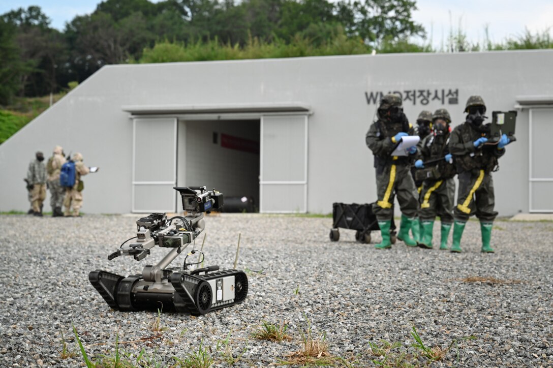 A bomb robot is rolling on the ground as four soldiers wearing camouflage military uniforms and personal protective equipment stand in the background holding laptops and other equipment. There is a large gray facility behind the soldiers.