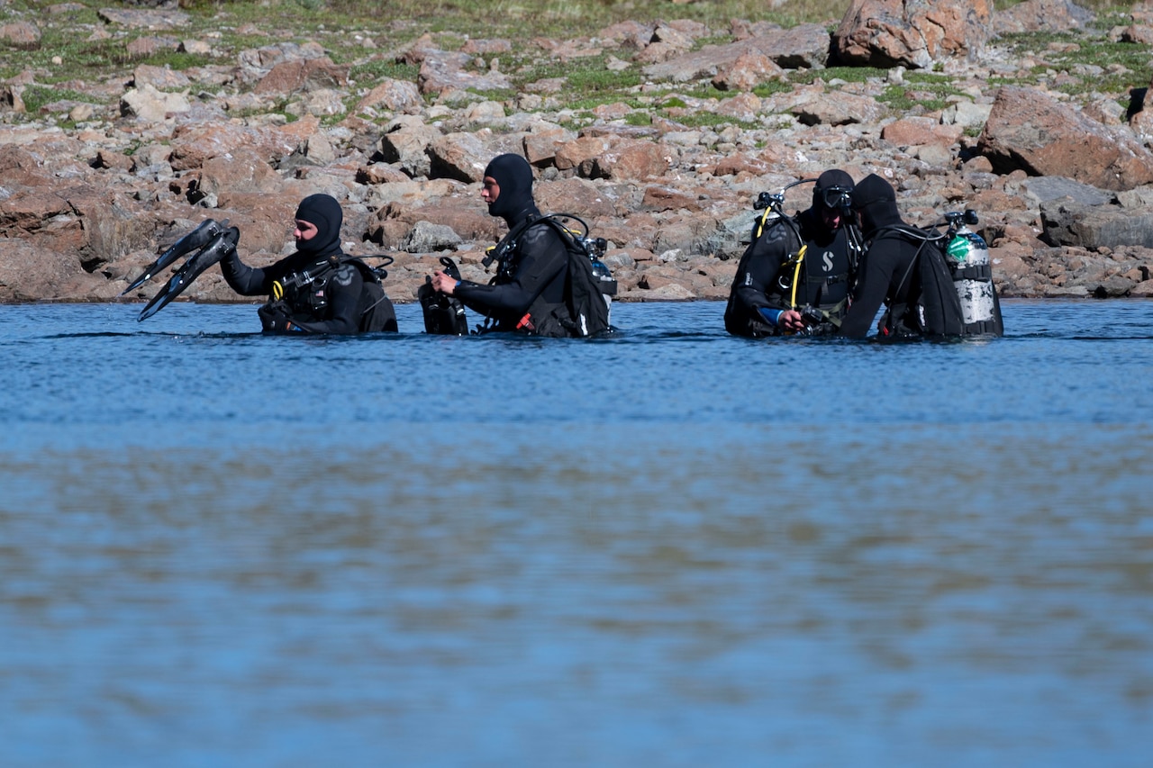 Four divers wearing wet suits and diving gear stand in waist-deep water. There are rocks on the bank in the background.