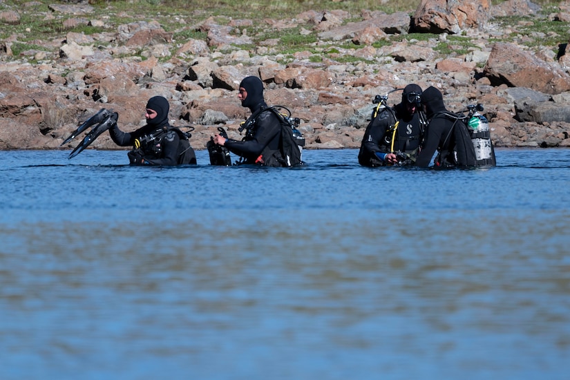 Four divers wearing wet suits and diving gear stand in waist-deep water. There are rocks on the bank in the background.