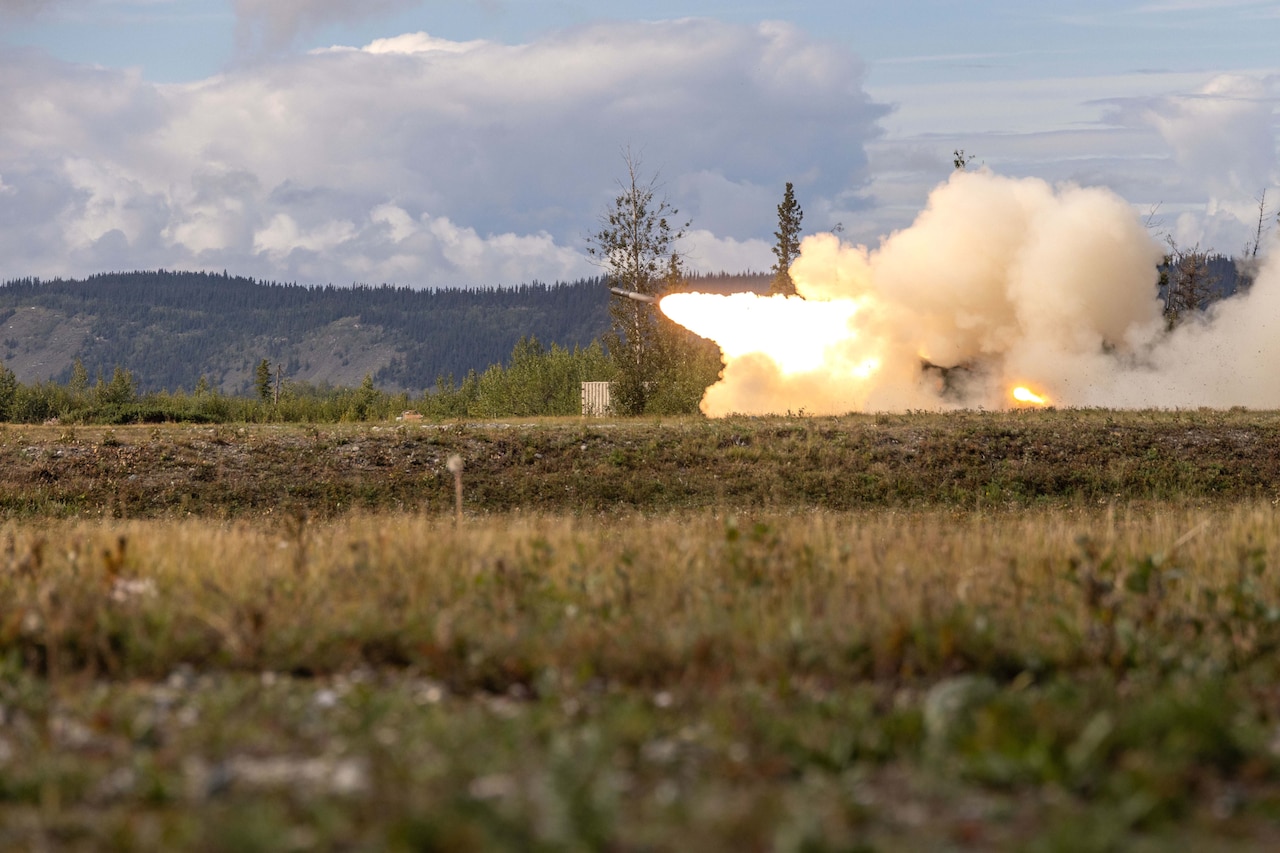 A rocket takes off from the ground as flames shoot out the bottom and create a large area of smoke. There is a field in the foreground and a hillside in the background.