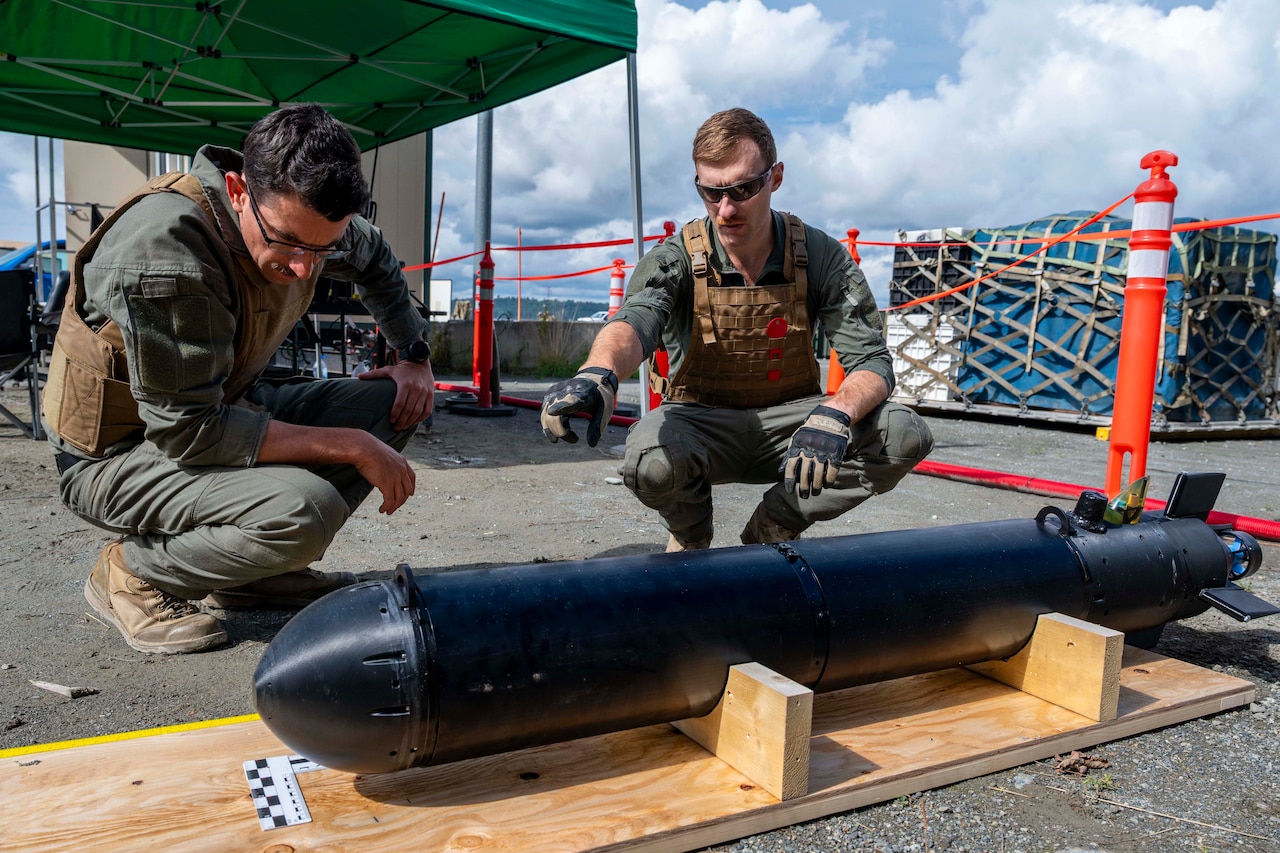 A man wearing a tactical military uniform, sunglasses and gloves points to the top of a torpedo while another man in similar attire looks at the torpedo.