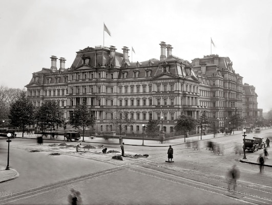 A black and white photograph of a gothic-style, five-story building with antique cars parked nearby.