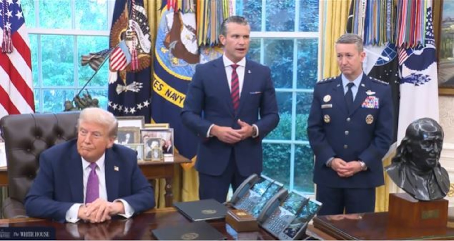 Three men, two in business attire and one in a dress military uniform, stand behind a large wooden desk with a window and a series of flags behind them. The man in the middle is speaking.