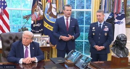 Three men, two in business attire and one in a dress military uniform, stand behind a large wooden desk with a window and a series of flags behind them. The man in the middle is speaking.