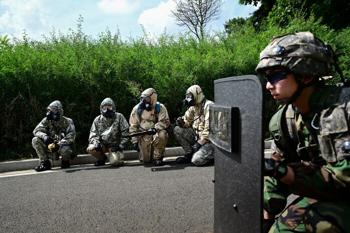A soldier wearing a camouflage military uniform holds a protective shield in front of their body while crouching on the ground. There are four soldiers in military camouflage uniforms and gas masks crouched un the background.