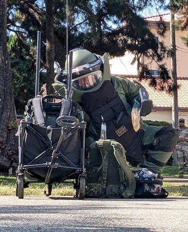 A soldier in a protective bomb suit crouches on the ground next to a backpack and roller cart. There are trees and a building in the background.