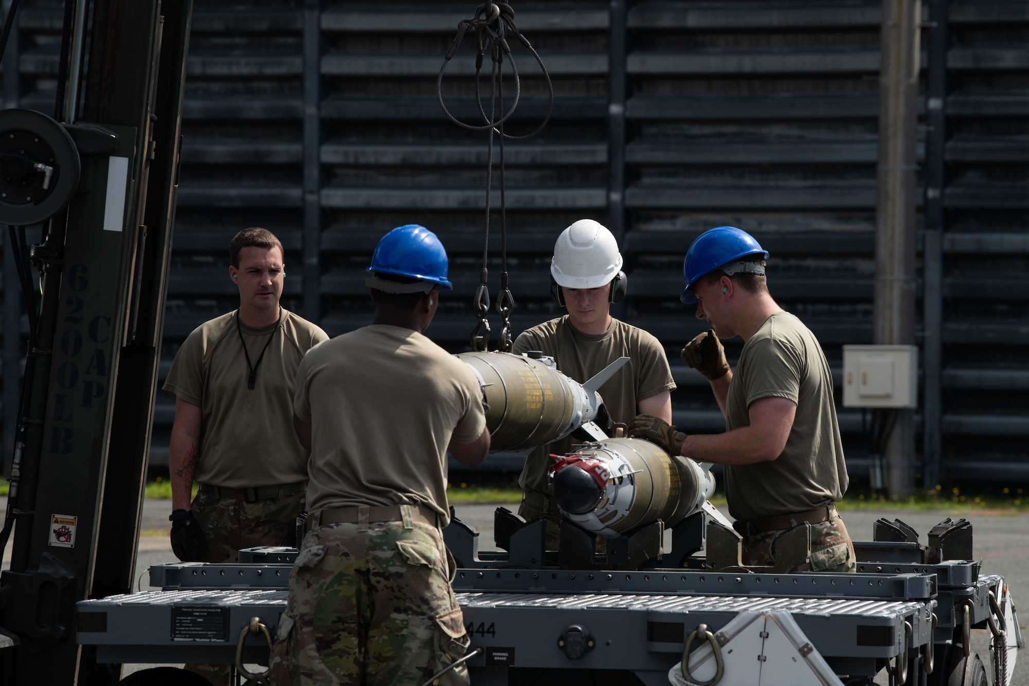 Uniformed men in hard hats lower a green bomb onto a gray cart with a forklift.