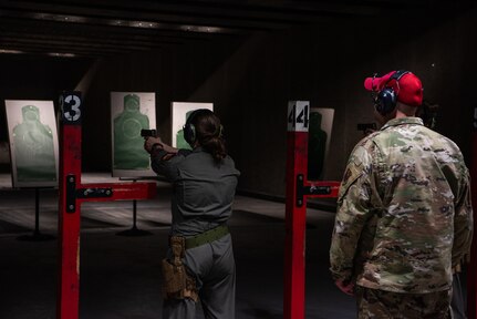 Uniformed man in red hat watches woman in green pilot jumpsuit aim a pistol at a target.