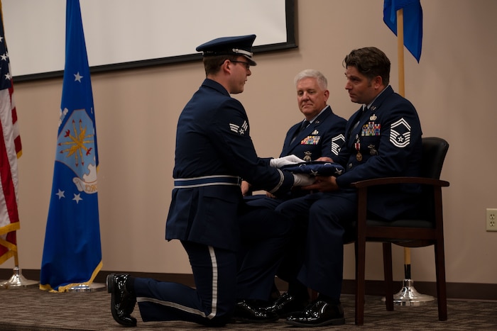 Sgt. Ganus sits in a chair as a member of the honor guard kneels before him and hands him a folded U.S. flag.