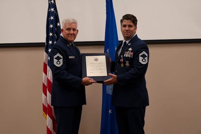 Sgt. Ganus and another Airmen hold on to a Certificate of Retirement and smile towards the camera.