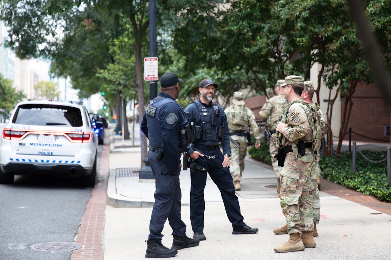 Two soldiers in military camouflage uniforms talk to two people in blue police uniforms as they stand on a sidewalk in a city. There is a police car parked on the street and two other soldiers in similar attire walking in the background.
