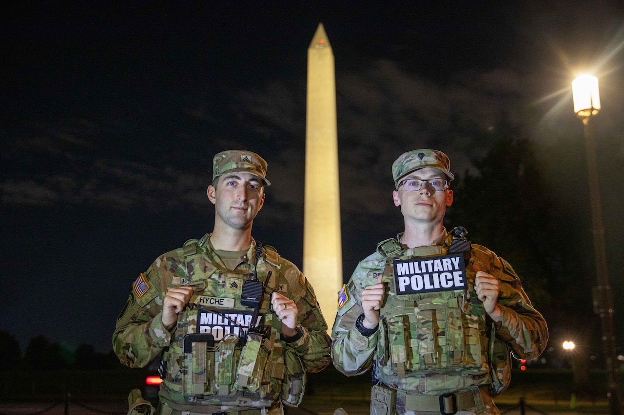 Two soldiers in camouflage military uniforms pose for a photo under a dark sky with the Washington Monument in the background. Both soldiers are wearing patches that read “military police.“