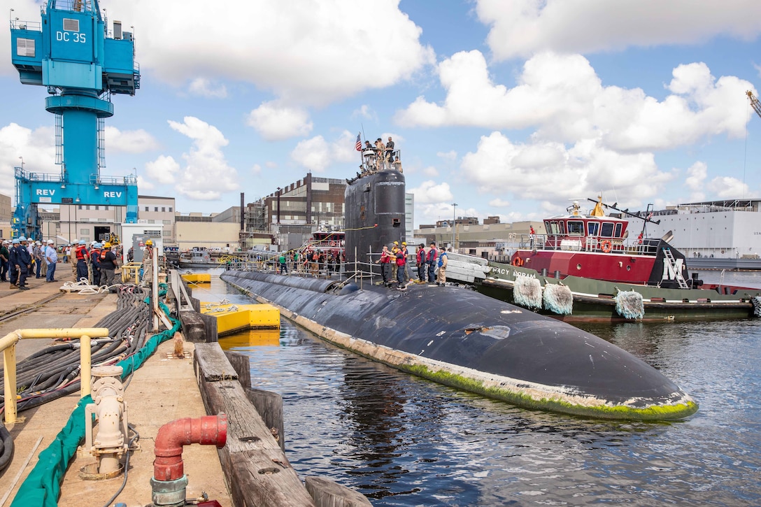Sailors stand on a submarine as it arrives at a port greeted by personnel wearing hard hats.
