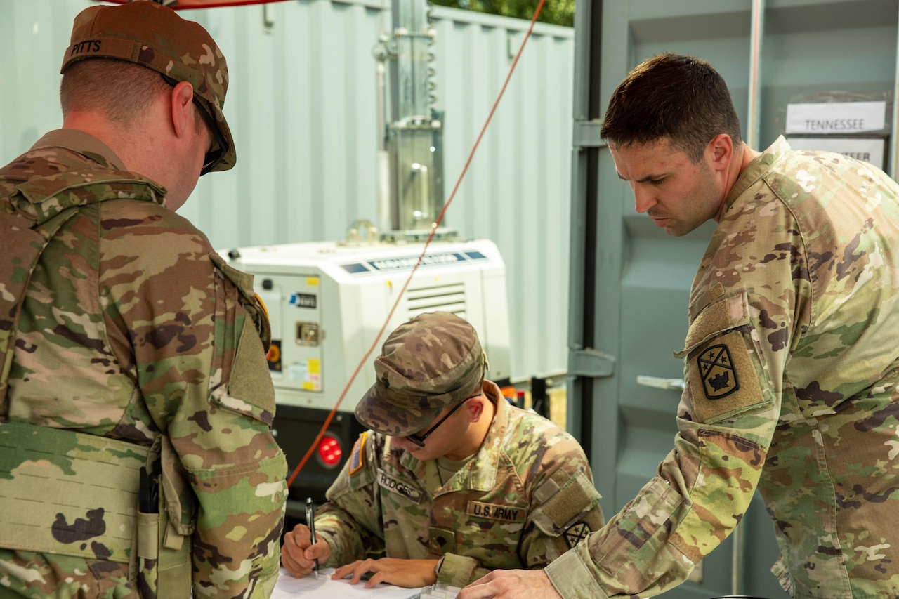 A soldier wearing a camouflage military uniform sifts through papers while another soldier in similar attire writes in a notebook while seated at a table. Another soldier in a camouflage uniform stands across the table looking down.