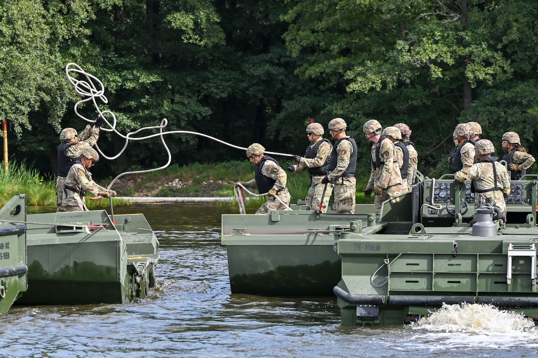 Soldiers in protective gear toss a line while riding in small boats near a wooded area.